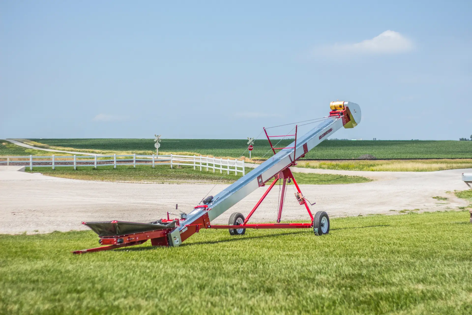 KSi Model 1212 Inclinator Series I conveyor on transport wheels at a farm site, red frame and galvanized cleated belt housing, KSi Conveyors branding visible
