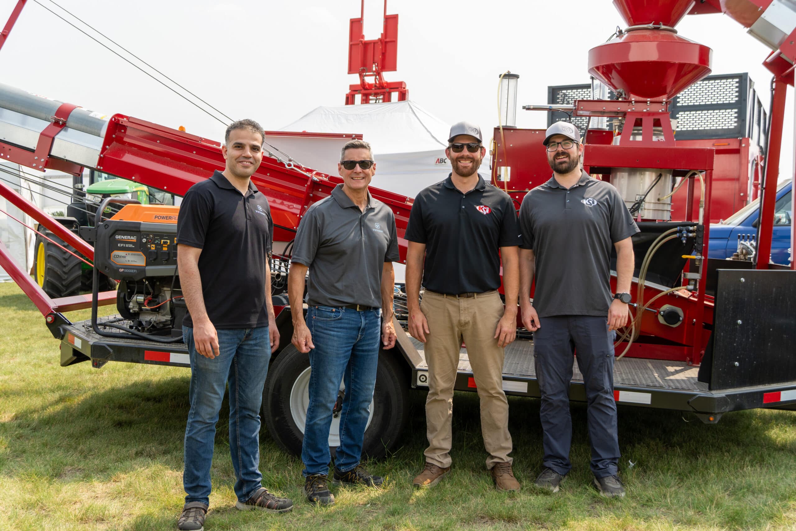 Vistasort's Steven Chivilo and Hamid standing with the KSi team in front of a red KSi seed treatment and conveyor system at Ag in Motion 2026 in Langham, Saskatchewan